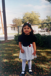 3 year-old Desiree wears a black top with a white skirt, white tights and black shoes. She has dark brown hair and bangs with a headband. She smiles big as she stands outside surrounded by green grass and red-tips. 